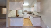 This single-floor residence offers a practical and comfortable layout with distinct living spaces. The kitchen is well-equipped, featuring white cabinetry with ample storage, dark countertops, and white appliances including a refrigerator, electric range, and dishwasher. A pass-through window connects the kitchen to the living area, creating an open feel between the two spaces. The kitchen also includes light hardwood-style flooring, adding warmth to the room.

The living area is generously sized, finished with wall-to-wall carpet in neutral tones and a textured ceiling. A baseboard heating unit runs along one wall, and a pass-through opening provides visual connectivity to adjacent spaces. The bedroom is a comfortable, carpeted room with a window that allows natural light and outdoor views, also equipped with baseboard heating.

The bathroom features light hardwood-style flooring, a single-sink vanity with white cabinetry and dark countertops, a large mirror, and a shower-tub combination with a decorative curtain. The overall aesthetic throughout the unit is clean and neutral, with warm beige and gray tones providing a cohesive, move-in-ready environment.