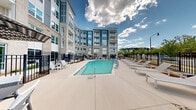 Located at 11201 Capital Blvd in Raleigh, NC, this outdoor amenity space is a well-appointed courtyard anchored by a sparkling resort-style swimming pool. The pool area is generously sized and surrounded by a broad concrete sundeck outfitted with rows of lounge chairs and shaded seating areas, providing ample space for relaxation and recreation. A gated black iron fence encloses the perimeter, ensuring privacy and security while maintaining an open, airy feel.

The courtyard is embraced on multiple sides by a striking multi-story building that combines classic brick masonry with modern design elements, including bold blue and gray paneling and expansive windows. Private balconies overlook the pool area from upper floors, creating a cohesive indoor-outdoor connection throughout the complex. Walkways with fresh landscaping, including mulched beds and young plantings, lead from building entrances directly to the pool deck.

Outdoor dining and gathering areas with umbrella-shaded tables are positioned around the deck, complementing the lounge seating. Views beyond the fenced boundary reveal a lush tree line, lending a natural backdrop to the space. Under clear North Carolina skies, this courtyard offers a bright, inviting atmosphere ideal for both quiet relaxation and social gatherings.