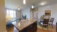 This single-floor, two-bedroom, two-bathroom residence offers a thoughtfully designed open-concept layout that seamlessly connects the kitchen, dining, and living areas. The kitchen serves as a focal point, featuring a large island with granite-style countertops, dark espresso cabinetry with brushed nickel hardware, and pendant lighting overhead that adds a refined touch. The flowing floor plan allows natural light from the living area windows to illuminate the entire shared space, creating a bright and welcoming ambiance. Hardwood-style flooring runs throughout the main living areas, lending warmth and continuity. The living room is spacious enough to accommodate comfortable seating arrangements, and a ceiling fan adds both style and practicality. A dedicated dining area sits adjacent to the kitchen, providing a natural transition between spaces. Neutral wall tones complement the darker cabinetry and flooring, creating a balanced, contemporary aesthetic. The layout is efficient and functional, offering a comfortable sense of space ideal for everyday living.
