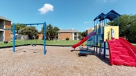 This community recreational space offers a well-equipped outdoor environment designed for active play and leisure. The playground area features a brightly colored multi-activity play structure complete with a red slide, climbing elements, and spring riders, complemented by a freestanding blue swing set. Wood chip surfacing surrounds the play equipment, providing a safety-conscious ground cover. The open green lawn extends beyond the playground, offering additional room for outdoor activities.

Adjacent to the playground sits a full-sized basketball court, enclosed by chain-link fencing for defined boundaries. The court features a red-painted key area, green playing surface, and a regulation-height hoop, making it suitable for organized games and casual play alike. Mature trees border the surrounding grounds, providing natural shade and a pleasant, park-like atmosphere under open skies.

The interior living space includes a spacious main living area with rich dark hardwood-style flooring, a textured ceiling, a ceiling fan with light fixture, and a wood-burning fireplace with a white surround. A sliding glass door allows natural light to fill the room and provides direct access to a private outdoor balcony. Neutral wall tones throughout contribute to a clean, versatile aesthetic suitable for a variety of interior styles.