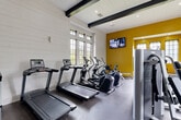 Located at 2400 E Nifong Blvd in Columbia, Missouri, this fitness center offers a comprehensive workout environment within a single, open floor layout. The space is thoughtfully designed, combining modern aesthetics with functional fitness infrastructure. Architecturally, the room features white shiplap walls, bold yellow accent walls, and striking exposed black ceiling beams that add an industrial-chic character. Recessed lighting and large multi-pane windows allow ample natural light to fill the space, creating an energizing atmosphere.

The cardio zone is well-appointed with multiple treadmills and elliptical machines positioned near the windows, offering outdoor views during workouts. A dedicated rowing machine and stationary bike round out the cardio offerings. For strength training, the facility includes a multi-station cable machine, a full rack of dumbbells ranging across various weights, medicine balls, kettlebells, exercise balls, and a punching bag — all neatly organized on a sturdy metal storage rack. An adjustable workout bench adds further versatility.

Wall-mounted flat-screen TVs are positioned throughout the space for entertainment. A built-in bench with cubbies near the entrance provides convenient storage. Durable rubber flooring covers the entire area, ensuring safety and comfort during exercise. Glass double doors serve as the main entry point, contributing to the open and accessible feel of this well-rounded fitness facility.