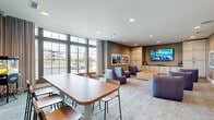 This well-appointed amenity lounge offers a thoughtfully designed single-floor layout that balances comfort, entertainment, and social functionality. The space is anchored by a media wall featuring a large flat-screen TV mounted above a run of built-in cabinetry with ample storage, dark countertops, and tall upper cabinets — ideal for hosting gatherings or casual viewing. A cluster of plush purple lounge chairs surrounds a central ottoman, creating an inviting conversation and entertainment area set atop neutral, patterned carpeting. Recessed lighting throughout the ceiling provides consistent, warm illumination across the space. Toward the front of the room, floor-to-ceiling multi-pane windows flood the interior with natural light and offer views of the surrounding exterior. A dining table with hairpin-style legs and warm wood chairs accommodates group seating near the windows. Decorative touches include a collection of colorful paddles mounted on the wall, a vintage-style popcorn machine, and a curated selection of framed artwork with nautical and coastal themes. Sheer floor-length curtains frame the windows, adding an elegant softness to the overall aesthetic. The combination of functional furnishings and cohesive coastal-inspired décor makes this lounge a versatile and welcoming communal space.