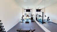 This fitness center is a thoughtfully designed exercise space that provides a comprehensive workout environment on a single level. The room features durable speckled rubber flooring throughout, offering both safety and comfort underfoot during workouts. Clean white walls and recessed ceiling lighting create a bright, energizing atmosphere, while a large wall-mounted mirror amplifies the sense of space and allows for proper form monitoring during exercise.

The cardio zone is anchored by a large picture window that frames views of the surrounding greenery, flooding the area with natural light. A treadmill, elliptical trainer, and recumbent stationary bike are positioned to take advantage of this outlook, with a ceiling-mounted flat-screen TV adding entertainment convenience. A stretch bench provides an additional functional element in this area.

The free weights section is well-stocked with a full dumbbell rack offering a range of weights, an adjustable workout bench, and an assortment of medicine and stability balls displayed on a dedicated stand. Stainless steel waste bins are strategically placed throughout the space for cleanliness. Access is provided through glass-panel doors, maintaining visual connectivity with adjacent areas. The overall design is clean, modern, and functional, making it an inviting space for a full range of fitness activities.
