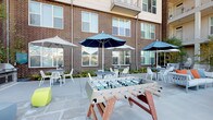 This outdoor lounge is a thoughtfully designed communal space nestled within a multi-story brick building complex, offering a dynamic blend of relaxation and recreation. The spacious patio is finished with large-format concrete pavers that create a clean, contemporary foundation throughout the area. Multiple distinct zones cater to a variety of activities: shaded dining tables with modern white chairs sit beneath striped and solid blue market umbrellas, providing comfortable spots for casual meals or conversation. Plush lounge seating with colorful accent pillows invites more leisurely gatherings, while vibrant geometric ottomans add a playful design touch. A foosball table serves as a central recreational feature, perfect for social engagement. Along one edge of the patio, a stainless steel grill station is set beside a masonry counter with a granite-topped surface, ideal for outdoor cooking and entertaining. Landscaped planting beds with ferns, ornamental shrubs, and young trees line the perimeter, softening the brick facades and adding natural texture. The space opens toward city views, with the surrounding urban skyline visible beyond a perimeter fence, creating a lively and connected outdoor atmosphere.