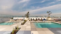 This expansive rooftop amenity deck offers a resort-inspired retreat elevated above the surrounding cityscape. At its heart is a generously sized swimming pool with crystal-clear turquoise water, complemented by a separate spa equipped with an accessibility lift, ensuring inclusive enjoyment for all. The pool deck is finished with large-format light-toned pavers that lend the space a clean, contemporary aesthetic.

Lush planted garden beds featuring drought-tolerant succulents and ornamental grasses are interspersed throughout the deck, adding a natural, textured contrast to the modern hardscape. Towering potted palms punctuate the space, enhancing its California resort atmosphere. Multiple clusters of woven rattan chaise lounges with teak frames are arranged to take full advantage of the abundant natural light and open-air environment.

A dedicated lounge area outfitted with cushioned daybeds and an outdoor entertainment setup — including a large flat-screen television, fireplace, and built-in grill station — provides a comfortable social hub. The perimeter is enclosed by seamless glass railings that preserve unobstructed panoramic views of the city skyline, with mountain ranges visible in the distance. The overall ambiance is sophisticated yet relaxed, capturing the essence of elevated urban outdoor living.