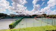 This single-floor residence is defined by its standout outdoor connection — a balcony enclosed by sliding glass doors that frame a direct view of the community's tennis courts below. The courts are well-maintained with hard green surfaces, surrounded by chain-link fencing and tall light poles for evening play. Lush green hedges and tropical landscaping border the complex, with neighboring residential buildings visible in the background beneath a dramatic Florida sky filled with rolling clouds. The sliding glass doors leading to the balcony allow generous natural light to filter into the interior, offering a seamless visual link between the indoor living space and the active outdoor amenities. The balcony itself features a railing that further enhances the open-air, resort-style feel of the setting. While several interior images are too dimly lit to reveal full detail, the overall character of the space speaks to a comfortable, amenity-rich residential environment. The combination of a private outdoor vantage point and access to recreational facilities such as the tennis courts contributes to an active and connected lifestyle, all within a well-established community setting defined by mature landscaping and a vibrant outdoor atmosphere.