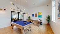 This single-level game room is a well-appointed recreational space designed for social entertainment. The interior is defined by white shiplap-clad walls that lend a clean, coastal aesthetic, complemented by light wood-tone flooring that runs throughout, creating a bright and cohesive environment. The room's generous footprint comfortably accommodates multiple game stations without feeling crowded.

At the center of the space sits a full-size pool table with a bold blue felt surface and a rustic wood frame, positioned beneath a striking mid-century modern chandelier with exposed Edison bulbs extending from angular black arms. A regulation-length shuffleboard table with a natural wood finish occupies an adjacent area, and a dartboard mounted on the wall adds another layer of entertainment options.

Natural light enters through large windows, offering views of the surrounding outdoor landscape, including palm trees, enhancing the room's airy atmosphere. Colorful framed artwork depicting aquatic and action-themed scenes adds vibrant personality to the walls. Yellow accent tables and potted tropical plants introduce playful pops of color. Large glass sliding doors connect the game room to an adjoining lounge area, promoting an open, flowing social environment.