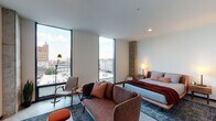 This single-floor studio is defined by its contemporary industrial aesthetic, seamlessly blending raw architectural elements with refined, modern furnishings. Polished concrete floors run throughout the entire space, providing a cohesive and sleek foundation that complements the clean white walls and exposed concrete columns — a standout structural feature that adds texture and character to the interior.

The main living area is bathed in natural light, courtesy of dramatic floor-to-ceiling windows that frame sweeping urban rooftop views and open sky. The space is thoughtfully arranged with a comfortable upholstered sofa, accent seating, a wall-mounted flat-screen television, and layered area rugs that soften the industrial finishes. A sliding barn door separates the living area from the kitchen, offering a practical yet stylish design detail.

The kitchen is compact and well-equipped, featuring warm walnut cabinetry, stainless steel appliances including a range and refrigerator, a tile backsplash, and a light countertop surface. A bold charcoal entry door anchors the kitchen entryway.

The bathroom showcases a distinctive graphic tile wall in dark tones with varied finishes, a glass-enclosed walk-in shower, a floating vanity with an illuminated mirror, and open wooden shelving stocked with linens — combining function with a refined, boutique-hotel aesthetic.