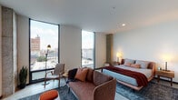 This single-floor studio is defined by its contemporary industrial aesthetic, seamlessly blending raw architectural elements with refined, modern furnishings. Polished concrete floors run throughout the entire space, providing a cohesive and sleek foundation that complements the clean white walls and exposed concrete columns — a standout structural feature that adds texture and character to the interior.

The main living area is bathed in natural light, courtesy of dramatic floor-to-ceiling windows that frame sweeping urban rooftop views and open sky. The space is thoughtfully arranged with a comfortable upholstered sofa, accent seating, a wall-mounted flat-screen television, and layered area rugs that soften the industrial finishes. A sliding barn door separates the living area from the kitchen, offering a practical yet stylish design detail.

The kitchen is compact and well-equipped, featuring warm walnut cabinetry, stainless steel appliances including a range and refrigerator, a tile backsplash, and a light countertop surface. A bold charcoal entry door anchors the kitchen entryway.

The bathroom showcases a distinctive graphic tile wall in dark tones with varied finishes, a glass-enclosed walk-in shower, a floating vanity with an illuminated mirror, and open wooden shelving stocked with linens — combining function with a refined, boutique-hotel aesthetic.