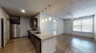 Located at 1719 Cable Ranch Rd in San Antonio, Texas, this single-story residence offers a well-designed open-concept layout that seamlessly connects the kitchen, dining, and living areas. The kitchen is a standout feature, showcasing dark espresso cabinetry with brushed nickel hardware, granite countertops with subtle veining, and a full suite of stainless steel appliances including a range, microwave, refrigerator, and dishwasher. A tile backsplash adds texture, while a peninsula with pendant lighting above creates a natural division between the kitchen and the open living space. Wood-style flooring extends throughout the main living areas, adding warmth and visual continuity, while crown molding contributes a refined architectural detail. The living area benefits from natural light through a front-facing window and is complemented by a ceiling fan. An arched doorway connects to additional rooms, adding a classic architectural touch. The bedroom features soft carpeting and a clean, neutral palette. The bathroom is appointed with large-format tile surrounding a soaking tub and shower combination, coordinating with the dark cabinetry above. The overall ambiance is modern and functional, with consistent finishes throughout.