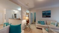 This single-floor residence at 2708 Salem Church Rd in Fredericksburg, Virginia, offers a well-organized layout connecting living, dining, kitchen, and bedroom spaces in a cohesive flow. The living room is finished with crown molding, neutral carpet, and a sliding glass door that draws in natural light while providing outdoor access. An open pass-through connects the living area to the kitchen, which features warm wood cabinetry, a breakfast bar counter, white appliances including a refrigerator, range, and microwave, and a conveniently tucked in-unit laundry closet behind louvered doors. The primary bedroom is generously proportioned, featuring a large window, carpet flooring, and a closet. A second bedroom offers similar comfort with its own window allowing natural light and views of surrounding greenery. Both bathrooms are functional and clean in design, each featuring a tub-shower combination and wood-look flooring with a vanity. A sitting area adjacent to the second bedroom provides additional flexible living space, accented by crown molding consistent with the main living area. Throughout, the unit maintains a calm, coastal-inspired palette with neutral walls and warm finishes.
