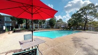 This inviting outdoor amenity space centers around a generously sized swimming pool with crystal-clear turquoise water, offering a refreshing retreat for residents and guests. The pool deck is finished in textured aggregate concrete, providing a safe and attractive surface that surrounds the pool on multiple sides. Poolside relaxation is made comfortable with the provision of lounge seating and umbrella-shaded tables, including a vibrant red patio umbrella that adds a pop of color to the setting. The area is enclosed by a wrought-iron perimeter fence, ensuring both safety and a sense of defined space. In the background, multi-story residential buildings with classic brick and light-colored facades are visible, along with exterior staircases that lend a traditional architectural character to the community. Mature trees frame the backdrop, offering natural shade and a lush, green ambiance that enhances the overall atmosphere. The space is bathed in natural sunlight under an open sky, making it a bright and welcoming environment. Steps and handrails provide easy pool access, while the well-maintained surroundings reflect a commitment to quality communal living.