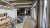 This single-floor communal space exudes a warm and inviting atmosphere, thoughtfully designed to balance comfort with sophistication. The open layout is anchored by a striking dark stone fireplace, which serves as the room's focal point and is complemented by a wall-mounted television above, making it an ideal gathering area for relaxation and socializing. Exposed wooden ceiling beams add a rustic yet refined architectural character to the space, contrasting beautifully with the smooth, neutral-toned walls and modern recessed lighting overhead. The flooring transitions seamlessly between warm hardwood and tile, delineating seating areas while maintaining a cohesive visual flow throughout. Plush teal accent chairs and tasteful area rugs provide comfortable, stylish seating arrangements that encourage conversation. Along one wall, a generous row of floor-to-ceiling French doors with elegant drapery panels floods the interior with abundant natural light while offering a sense of openness and connection to the outdoors. In the background, a dining or bar area with pendant lighting is visible, extending the functionality of the space. Overall, this communal area offers a refined, resort-inspired environment well-suited for both quiet retreats and social gatherings.