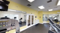This fitness center is a thoughtfully designed, single-level workout facility offering a well-rounded selection of exercise equipment. The space is divided into distinct zones, connected by elegant white French doors with glass panes that allow light and sightlines to flow between areas. The walls are painted in a warm yellow accent tone complemented by crisp white trim and neutral gray ceilings, creating an energetic yet welcoming atmosphere.

Flooring throughout is dark rubber, providing durability and comfort underfoot. Large wall-mounted mirrors visually expand the space and allow users to monitor their form during workouts. Flush-panel and recessed lighting fixtures illuminate the interior evenly, while multiple windows along the perimeter bring in natural light and offer views of the surrounding greenery.

The cardio section is equipped with treadmills, elliptical machines, and stationary bikes, while the strength training area features cable and resistance machines. A wall-mounted television is positioned for viewing during workouts. Built-in white shelving units provide storage for personal items or fitness accessories. An emergency phone and safety signage are also present, reflecting attention to user safety throughout the facility.