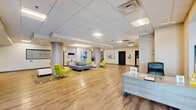 Located at 3801 Conshohocken Ave in Philadelphia, PA, this single-floor commercial space offers a generously sized, open layout designed for a professional and welcoming environment. The expansive lobby serves as the centerpiece, featuring warm hardwood-style flooring, a drop ceiling with recessed panel lighting, and elegant cylindrical columns with textured finishes that define the space architecturally. A dedicated reception desk area anchors the entry point, providing a clear focal area for visitors upon arrival. The seating lounge within the lobby is thoughtfully arranged with contemporary blue and vibrant chartreuse accent chairs, complemented by a patterned area rug that adds visual warmth. Abstract artwork in cool blue and neutral tones is displayed throughout, lending a polished, curated aesthetic to the walls. A sculptural metallic art piece near the entryway adds a distinctive decorative touch. The hallways extend from the main lobby and are equally well-appointed, featuring the same light hardwood flooring, clean white walls adorned with framed artwork, and recessed ceiling lighting. Elevator access and stairwell connections are visible within the corridor areas. Large windows allow natural light to filter in, enhancing the bright, airy ambiance throughout the entire floor.