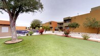 Located at 1520 University Blvd NE in Albuquerque, New Mexico, this single-story property showcases classic Southwestern adobe-inspired architecture with warm earth-tone stucco exteriors in shades of tan and terracotta. The building's clean, flat-roofed design is complemented by accents of bold orange on doorways and trim, reflecting the region's distinctive architectural identity.

At the heart of the property lies an expansive, well-maintained communal courtyard that serves as a central gathering space. A generous stretch of lush green lawn provides an open, inviting atmosphere, bordered by mature shade trees, ornamental grasses, flowering shrubs, and decorative rock landscaping that adds textural variety throughout the grounds. A large, established shade tree anchors the courtyard, surrounded by a circular planting bed.

Outdoor seating areas are thoughtfully distributed across the courtyard, including colorful Adirondack chairs and patio furniture with tables and chairs, offering residents multiple spots to relax and socialize. A grill station is also present, enhancing the outdoor living experience. Individual unit entrances face directly onto the courtyard, each with a covered walkway, creating a connected and community-oriented layout. The surrounding multi-story buildings feature balconies and staircases that overlook the landscaped grounds, completing this cohesive, pedestrian-friendly Albuquerque property.