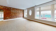 Located at 846 Main St in downtown Buffalo, NY, this one-bedroom, one-bathroom residence blends industrial character with modern updates across a well-appointed single floor. The open-concept kitchen and living area serves as the heart of the unit, where exposed red brick walls create a striking architectural focal point. The kitchen is equipped with stainless steel appliances, including a gas range, microwave, and French-door refrigerator, complemented by dark granite countertops, light maple cabinetry, and a central island with built-in storage. Stone tile flooring grounds the space and flows through the kitchen and entry areas. The spacious living room is bathed in natural light from multiple large windows, including a distinctive oval porthole window, offering views of the surrounding Buffalo streetscape. Plush neutral carpeting extends throughout the living area, hallway, and bedroom, adding warmth and comfort. The bedroom is generously sized, featuring multiple windows that frame views of neighboring greenery and the city skyline. The bathroom offers a clean, modern aesthetic with large-format white tile flooring, a wood-toned vanity with dark granite, and a frosted window for natural light and privacy. A utility room, closet, and hallway complete the layout, providing practical everyday storage and functionality.