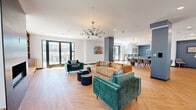 Located at 201 Munson Street in New Haven, CT, this single-floor combined kitchen and living space offers a generous, open-concept layout with thoughtful modern design throughout. Herringbone-patterned hardwood floors flow seamlessly across the entire space, lending warmth and visual interest underfoot.

The expansive living area is anchored by a sleek, tile-surround fireplace and flanked by built-in wooden shelving and cabinetry with integrated window bench seating. Large windows and floor-to-ceiling glass sliding doors flood the room with natural light and provide direct access to an outdoor terrace, offering treetop views. A striking multi-arm chandelier with teal glass globes serves as a stylish focal point overhead.

The kitchen is a standout feature, boasting bold navy blue cabinetry paired with quartz countertops and stainless steel appliances, including a large refrigerator and a professional-grade gas cooktop beneath a substantial stainless steel range hood. A generously sized island with bar seating provides both prep space and casual dining. Open shelving, a white tile backsplash, and curated décor complete the kitchen's contemporary aesthetic.

The overall ambiance is bright, airy, and sophisticated, blending mid-century modern furnishings with clean contemporary architecture for a welcoming and functional communal environment.