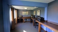This single-floor business center is designed to provide a functional and comfortable workspace for productivity. The room is finished with cool blue-grey walls that create a calm, professional atmosphere, complemented by neutral-toned carpeting underfoot. Recessed ceiling lighting, along with a central flush-mount fixture, ensures the space is well-lit throughout the day, while a window fitted with warm curtain panels allows for natural light to filter in.

The workstations are arranged along built-in wooden desks that line two walls, offering ample surface space for multiple users at once. Each station is equipped with computers, and a shared printer is conveniently accessible at the end of one desk run. Sturdy dark wood chairs with cushioned seats provide comfortable seating at each workstation.

Decorative framed artwork adorns the walls, adding a polished and welcoming touch to the utilitarian setting. A potted plant near the desks brings a subtle natural element into the room. The overall layout is thoughtfully organized to maximize usability while maintaining an open, uncluttered feel — making this business center a practical and inviting space for work and administrative needs.