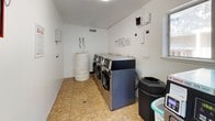 This dedicated laundry facility is designed for practical, high-capacity use. The room is finished with clean white walls and durable tile flooring, creating a bright and functional environment. At its core, the space is outfitted with multiple commercial-grade front-loading washers and dryers from the WASH WBI series, including Speed Queen commercial machines, all mounted on pedestals for added convenience. The machines feature digital control panels, card/coin payment systems, and are arranged along the walls to maximize usable floor space. Instructional and operational signage is posted on the wall above the machines for easy reference. A large insulated water tank with visible plumbing connections is situated in the corner of the room, supporting the facility's water supply needs. Additional utility components, including control panels and electrical fixtures, are mounted on the walls. The room also includes a stacked washer-dryer unit, offering further laundry capacity. Natural light filters in through windows on one wall, and a dedicated entry door provides direct access to the facility. A waste bin and laundry basket are conveniently placed within the space, completing this fully operational, shared laundry room.