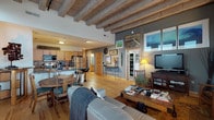 This single-floor space showcases a compelling loft-inspired design highlighted by striking exposed wooden beam ceilings that run throughout the main living areas, lending an industrial yet warm character to the interior. Light hardwood floors extend across the open-concept layout, seamlessly connecting the living, dining, and kitchen areas into one cohesive space.

The kitchen is well-appointed with stainless steel appliances, including a refrigerator and range, complemented by light wood cabinetry, granite-style countertops, and a tiled backsplash. A breakfast bar with modern stools provides casual seating. The adjacent dining area comfortably accommodates a full table and chairs, while the living room features large windows that invite natural light and offer urban views of the surrounding neighborhood.

The primary bedroom continues the exposed beam ceiling aesthetic with soft carpeting, grey accent walls, and dual windows with city views. A versatile den or office space offers carpeted flooring, a large window, and ample wall space for artwork. Two bathrooms serve the residence: one features a shower-tub combination with tile flooring, while the other offers a spacious vanity with dual mirrors and a separate glass-enclosed shower. Throughout the space, curated artwork and warm lighting contribute to an eclectic, lived-in ambiance.