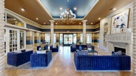 Located in Fort Worth, TX, this clubhouse at Lake Pointe offers a sophisticated and welcoming environment designed for community gatherings and relaxation. The grand living room immediately commands attention with its soaring ceilings, dramatic tray detail painted in deep blue and gold tones, and striking horizontal striped wall design in navy, tan, and warm brown. Towering twisted stone columns anchor the space, complementing a stately decorative fireplace framed by built-in shelving and elegant artwork. Plush royal blue velvet sofas are arranged in multiple seating groupings atop large area rugs over polished tile flooring, while a cascading gold chandelier adds a refined focal point overhead. French doors and expansive windows flood the space with natural light and offer views of the surrounding landscaped grounds. The dining and kitchen area is well-appointed with rich wood cabinetry, dark granite countertops, an arabesque tile backsplash, and stainless steel appliances including a full-size refrigerator. A cozy nook with large windows and wicker seating provides a casual spot to relax. Throughout the hallway and transition spaces, arched windows and doorways maintain the architectural elegance that defines this impressive clubhouse.