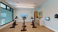 Located at 2260 Ringling Blvd in Sarasota, Florida, this single-floor fitness studio offers a well-appointed, purpose-built environment designed to support a variety of workout routines. The space is organized into distinct functional zones, seamlessly connected by light wood-style flooring and softly painted walls in a calming sage-green and neutral palette.

The cycling area is equipped with multiple stationary spin bikes arranged in a row before a large wall-mounted flat-screen display, ideal for guided or virtual cycling classes. Built-in wooden cabinetry and shelving provide convenient storage, while framed artwork adds a refined, welcoming touch to the workout environment.

Adjacent to the cycling zone, a spacious open training area features floor-to-ceiling mirrors along one wall, amplifying the sense of space and allowing users to monitor their form. Large dark-framed windows flood the room with natural light and offer views of the surrounding outdoor area, including a glimpse of a pool. A Life Fitness equipment rack is stocked with stability balls, kettlebells, resistance tools, and exercise mats, supporting strength and functional training.

The studio is accessed through glass-paneled double doors and includes a digital access panel and posted guidelines, reflecting thoughtful facility management. Linear LED lighting throughout ensures consistent, bright illumination across the entire space.