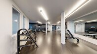 This fitness center offers a comprehensive, single-floor workout facility designed with both functionality and aesthetics in mind. The space features a clean, modern aesthetic characterized by a neutral white and dark charcoal color palette, durable dark rubberized flooring, and bright linear LED lighting that ensures excellent visibility throughout.

The cardio zone is equipped with multiple treadmills and upright exercise bikes, each featuring integrated display screens. A dedicated strength training area houses a multi-function cable machine, adjustable benches, and a full rack of dumbbells alongside kettlebells, catering to a wide range of training styles. Structural columns are integrated neatly into the floor plan, helping to define distinct workout zones without interrupting the overall open feel of the space.

Full-length mirrors span one wall, enhancing the sense of space and allowing users to monitor their form during exercise. Large windows along the perimeter flood the facility with natural light and provide views of the surrounding exterior. Additional amenities include a wall-mounted split air conditioning unit, a dual-spout water fountain station, a built-in wooden storage shelving unit, and a dedicated entry with a glass-paneled black door. The overall ambiance is that of a well-appointed, professional-grade fitness facility.