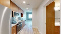 This single-floor unit showcases a compelling blend of industrial and contemporary design elements throughout its well-organized layout. The kitchen is a standout feature, equipped with stainless steel appliances including a side-by-side refrigerator with ice maker, electric range, over-the-range microwave, and dishwasher. Rich warm-toned wood cabinetry lines the walls floor-to-ceiling, complemented by a sleek subway tile backsplash in a soft blue-gray hue and light-colored countertops. Light wood-look flooring flows consistently throughout the entire unit, creating visual continuity.

The living area and bedroom share a striking design detail: exposed raw concrete walls that lend an authentic industrial character to the space. Windows along the concrete walls invite natural light and provide open views of the surrounding cityscape. A ceiling fan is installed in the bedroom, enhancing comfort. The bathroom features a wood-toned vanity with a crisp white countertop and undermount sink, a large mirror, and a tub-shower combination with vertically laid stone-look tile surround. Clean white walls and recessed lighting throughout ensure the space feels bright and polished.