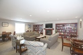 This single-floor space is designed to foster comfort and community, beginning with a well-appointed lobby entrance. The entry area features carpet flooring, recessed lighting, a welcome station, and large glass double doors that allow natural light to filter in. A rich wood interior door and wall-mounted signage add a polished, organized feel to the arrival experience.

The main lounge area serves as the heart of the space, offering generous room for gathering and relaxation. Plush sofas and accent chairs in neutral tones are arranged around a central coffee table, creating multiple inviting conversation areas. A gas fireplace with a warm wood mantel anchors one wall, flanked by floor-to-ceiling built-in bookshelves stocked with reading material. A wall-mounted flat-screen television sits above the fireplace, adding a functional entertainment element.

Recessed lighting throughout provides consistent, even illumination, complemented by natural light from windows along the perimeter. A small dining area with dark wood table and chairs is tucked nearby, offering additional functional space. Framed artwork and decorative accents contribute to the warm, residential ambiance of the overall environment.
