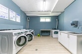Located at 446 Kerr St in Oakville, Ontario, this shared laundry facility is a well-organized and functional amenity space. The room is finished with cool blue-grey walls, light wood-look flooring, and a decorative textured ceiling that adds a touch of character. Fluorescent overhead lighting ensures the space is well-lit throughout.

The main area is equipped with multiple front-loading Maytag washers and dryers, arranged along the wall for efficient use of the space. A dedicated dryer alcove features an additional top-load dryer with a vented exhaust duct, accompanied by a built-in white folding counter — ideal for sorting and folding laundry. A small window in this alcove allows for natural light and ventilation.

Near the entrance, a stainless steel utility sink with white cabinetry provides added convenience. A digital payment kiosk is positioned nearby, offering a coin-free payment option for users. Informational signage and laundry instructions are clearly posted on the walls for easy reference. A seating chair is available for those waiting. The space is thoughtfully designed to be clean, practical, and easy to navigate, serving as a convenient shared resource for residents of the building.