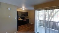 This single-floor residence offers a thoughtfully designed layout that balances functionality and style. The kitchen is a standout feature, showcasing rich dark cabinetry that extends to the ceiling, complemented by a decorative stone tile backsplash that adds texture and visual interest. Black appliances, including a refrigerator, stove, and microwave, lend a sleek, cohesive look to the space. Light-toned countertops provide a pleasing contrast against the darker elements, creating a balanced aesthetic. The kitchen flows seamlessly into the adjoining living area, where warm hardwood-style flooring transitions to soft carpeting, defining the separate zones naturally. Neutral, earthy wall tones throughout create a warm and inviting ambiance. A set of vertical blinds lines a full-length glass door, allowing abundant natural light to filter into the space while offering easy access to an outdoor area. The exterior view visible through the door suggests a pleasant outdoor setting. Overhead, a flush-mount ceiling light provides ample illumination, and the textured ceiling adds a subtle layer of detail to the overall design. This residence presents a comfortable and practical living environment with appealing finishes throughout.