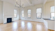 This thoughtfully designed single-floor unit blends classic architectural character with modern finishes throughout its well-appointed layout. Soaring ceilings amplify the sense of openness across the living spaces, which are bathed in natural light from a generous array of tall, black-framed windows offering views of the surrounding urban streetscape.

The expansive combined living and dining area is anchored by a striking decorative fireplace with an arched black surround, flanked symmetrically by windows on either side. Light-toned hardwood floors run continuously throughout, enhancing the bright, airy ambiance. An elegant geometric chandelier and exposed ductwork add an industrial-chic touch overhead.

The kitchen is a standout feature, showcasing white shaker cabinetry, classic white subway tile backsplash, dark stone countertops, a central island with matching stone surface, and a full suite of stainless steel appliances. Pendant globe and sconce lighting complete the refined aesthetic.

A separate living room area benefits from additional windows and a ceiling fan, maintaining the unit's consistent brightness and airflow. The bathroom is finished with penny round tile flooring, a teal vanity with stone countertop, and a glass-enclosed shower, offering a clean, contemporary feel throughout.