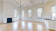 This thoughtfully designed single-floor unit blends classic architectural character with modern finishes throughout its well-appointed layout. Soaring ceilings amplify the sense of openness across the living spaces, which are bathed in natural light from a generous array of tall, black-framed windows offering views of the surrounding urban streetscape.

The expansive combined living and dining area is anchored by a striking decorative fireplace with an arched black surround, flanked symmetrically by windows on either side. Light-toned hardwood floors run continuously throughout, enhancing the bright, airy ambiance. An elegant geometric chandelier and exposed ductwork add an industrial-chic touch overhead.

The kitchen is a standout feature, showcasing white shaker cabinetry, classic white subway tile backsplash, dark stone countertops, a central island with matching stone surface, and a full suite of stainless steel appliances. Pendant globe and sconce lighting complete the refined aesthetic.

A separate living room area benefits from additional windows and a ceiling fan, maintaining the unit's consistent brightness and airflow. The bathroom is finished with penny round tile flooring, a teal vanity with stone countertop, and a glass-enclosed shower, offering a clean, contemporary feel throughout.