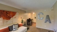 This single-floor residence offers a welcoming and cohesive layout that blends the living, dining, and kitchen areas into one harmonious open-concept space. The living room is warmly appointed with soft, neutral-toned walls and plush carpeted flooring, creating a comfortable and inviting atmosphere. A bold abstract artwork anchors one wall, complemented by an industrial-style wall-mounted lamp that adds a modern edge to the cozy setting. The space flows naturally into the dining area, where a dark bistro-style high table and bar stools serve as a functional yet stylish centerpiece. Beyond the dining area, the kitchen is visible through an open passageway, featuring white cabinetry and warm lighting that contribute to the home's bright and airy feel. The textured ceiling throughout adds a classic residential character to the space. Thoughtful decorative touches — including a large metal letter accent on the wall and curated accessories — give the home a personalized charm. The seamless connection between living, dining, and kitchen areas makes this space ideal for both relaxed everyday living and casual entertaining.