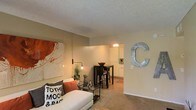 This single-floor residence offers a welcoming and cohesive layout that blends the living, dining, and kitchen areas into one harmonious open-concept space. The living room is warmly appointed with soft, neutral-toned walls and plush carpeted flooring, creating a comfortable and inviting atmosphere. A bold abstract artwork anchors one wall, complemented by an industrial-style wall-mounted lamp that adds a modern edge to the cozy setting. The space flows naturally into the dining area, where a dark bistro-style high table and bar stools serve as a functional yet stylish centerpiece. Beyond the dining area, the kitchen is visible through an open passageway, featuring white cabinetry and warm lighting that contribute to the home's bright and airy feel. The textured ceiling throughout adds a classic residential character to the space. Thoughtful decorative touches — including a large metal letter accent on the wall and curated accessories — give the home a personalized charm. The seamless connection between living, dining, and kitchen areas makes this space ideal for both relaxed everyday living and casual entertaining.