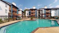 This expansive outdoor amenity space centers around a generously sized resort-style swimming pool with clear turquoise water, framed by brick-accented coping and decorative landscaping islands featuring palm plants and river rock. The pool area is enclosed by a wrought iron fence with ornamental detailing, providing both security and an elegant aesthetic.

The surrounding multi-story residential buildings display a bold contemporary design, combining white stucco exteriors with teal-painted brick columns and warm wood-toned horizontal accent panels on the balconies. Chimneys punctuate the roofline, adding architectural character throughout the complex.

The pool deck offers ample room for lounging, with multiple cushioned chaise lounges arranged beneath vibrant orange market umbrellas that provide shade on sunny days. A dedicated lounge zone features a covered pergola with a wood-slat ceiling, complemented by wicker egg chairs and a communal seating arrangement around a fire pit-style feature, creating an inviting social atmosphere.

Decorative rock gardens border the fenced perimeter, adding low-maintenance greenery that enhances the overall outdoor aesthetic. The entire space is bathed in natural sunlight, making it an ideal setting for relaxation and recreation. The cohesive color palette of teal, white, orange, and warm wood tones gives the pool area a lively yet polished resort-like character.