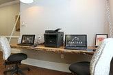 This single-floor community or leasing office for Panther Residential Management offers a welcoming and well-appointed environment designed with both style and practicality in mind. The space features cork flooring throughout, lending a warm, natural feel underfoot that complements the overall aesthetic. A standout business center workstation is anchored by a live-edge wood floating desk, suspended by industrial chain hardware and illuminated by a pendant light fixture above — a distinctive design detail that blends rustic and modern elements. The workstation is equipped with computers and a multifunction printer, providing residents or guests with convenient office resources. Decorative rustic artwork, including a wooden horse panel, adds character and charm to the walls. The kitchenette or lounge area features dark wood cabinetry, black granite countertops, a stainless steel refrigerator, and a row of industrial-style barstools along a peninsula counter, ideal for casual gatherings. Warm pendant lighting illuminates this area, enhancing the inviting atmosphere. Natural light filters in through large windows dressed with white horizontal blinds, offering views of lush greenery outside. A decorative glass panel featuring an elegant reed or branch motif adds an artistic touch, serving as a refined architectural accent within the space.