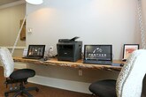 This single-floor community or leasing office for Panther Residential Management offers a welcoming and well-appointed environment designed with both style and practicality in mind. The space features cork flooring throughout, lending a warm, natural feel underfoot that complements the overall aesthetic. A standout business center workstation is anchored by a live-edge wood floating desk, suspended by industrial chain hardware and illuminated by a pendant light fixture above — a distinctive design detail that blends rustic and modern elements. The workstation is equipped with computers and a multifunction printer, providing residents or guests with convenient office resources. Decorative rustic artwork, including a wooden horse panel, adds character and charm to the walls. The kitchenette or lounge area features dark wood cabinetry, black granite countertops, a stainless steel refrigerator, and a row of industrial-style barstools along a peninsula counter, ideal for casual gatherings. Warm pendant lighting illuminates this area, enhancing the inviting atmosphere. Natural light filters in through large windows dressed with white horizontal blinds, offering views of lush greenery outside. A decorative glass panel featuring an elegant reed or branch motif adds an artistic touch, serving as a refined architectural accent within the space.