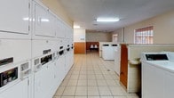 This single-floor laundromat offers a practical and functional layout designed to accommodate a high volume of users efficiently. The space is characterized by durable, light-colored tile flooring throughout and two-tone walls featuring a warm terracotta orange lower half paired with a cream upper half, giving the facility a clean and identifiable aesthetic.

The main area houses a substantial row of front-load washing machines and dryers lining one wall, each equipped with individual digital payment panels for convenient, cashless operation. Adjacent to the machines, a long folding counter provides ample workspace for sorting and folding laundry. A prominent bank of white storage lockers offers secure space for patrons to store personal belongings during their visit.

A designated waiting area features a small row of white molded chairs, providing a comfortable spot for customers to relax while their laundry cycles complete. Overhead fluorescent lighting ensures the entire space is well-lit, and ventilation is provided through ceiling-mounted HVAC units. A large utility waste bin is positioned for easy access, supporting cleanliness and day-to-day operations.

Natural light filters in through barred windows, and a standard entry door provides straightforward access. Informational signage, including laundry instructions from ARN Laundry Co., is posted on the walls for patron guidance. Overall, the facility is designed with practicality, durability, and user convenience at the forefront.