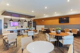 Located at 151 Integra Vistas Dr in Chattanooga, Tennessee, this single-floor game room is thoughtfully designed as a dedicated entertainment and social space. At its center sits a full-size round poker table surrounded by rich dark wood chairs with tan leather upholstered seats, serving as the focal point of the room. The space is anchored by a built-in wet bar niche, framed by a striking textured mosaic tile accent wall in dark metallic tones, complemented by warm amber-hued wall panels on either side. Flanking the wet bar are two built-in bench seating areas with cushioned pads and decorative pillows, ideal for casual conversation or spectating. Recessed ceiling lighting and pendant fixtures cast a warm, inviting glow throughout. On the opposing wall, a wall-mounted flat-screen television and an electronic dartboard with a chalkboard scoreboard provide additional entertainment options. A row of clerestory windows near the ceiling introduces natural light without compromising wall space. The flooring features a neutral-toned patterned carpet that adds texture and warmth underfoot. Abstract artwork decorates the walls, lending a contemporary art-gallery feel. Overall, this game room balances a sophisticated aesthetic with highly functional amenities, making it a premier space for social gatherings and recreational activities.