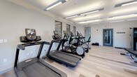Located at 100 Gateway in Scarborough, Maine, this modern fitness center is designed to support a comprehensive workout experience across multiple dedicated zones. The space features light-toned luxury vinyl plank flooring throughout, lending a clean and contemporary aesthetic that complements the bright, neutral walls and recessed linear lighting overhead.

The cardio area is well-appointed with a lineup of treadmills, elliptical trainers, and a stationary bike, all positioned alongside large mirrors that expand the sense of space and allow users to monitor their form. Natural light filters in through multiple windows, creating an open and energizing atmosphere.

A dedicated strength training zone includes a functional cable machine and adjustable weight benches, positioned near a large window that offers views of the surrounding area. Wall-mounted hooks provide convenient storage for accessories and attachments.

A separate studio and flexibility room rounds out the facility, featuring wide-plank flooring in a contrasting tone and an organized rack stocked with resistance bands, exercise balls, medicine balls, and yoga mats. Ample natural light pours through double-hung windows, making this an ideal space for stretching, yoga, or group exercise. A decorative textured wall panel adds a distinctive design touch to the room.
