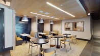 Located at 3560 Rambla Place in Santa Clara, California, this single-floor commercial space offers a thoughtfully designed environment that balances casual dining, collaborative work, and quiet retreat. The layout is divided into distinct yet interconnected zones, each with its own character and function.

The dining area is defined by warm natural wood slat ceilings, linear pendant lighting, and textured deep-navy brick accent walls. Booth seating with mustard-toned cushions lines the walls, complemented by round black bistro tables and light natural wood chairs arranged throughout. Abstract artwork in earth tones adds visual interest to the walls.

Transitioning through the space, large-format dark stone-look tile flooring anchors a corridor and transition zone featuring exposed industrial elements — dark-painted ductwork and structural columns — that contrast with the warmer finishes nearby. A glass-paneled partition separates this area from an adjacent lounge, allowing views while maintaining definition.

The open lounge area offers flexible seating arrangements, including freestanding acoustic privacy panels, round tables, and accent chairs in natural wood and yellow upholstery. A large window at the far end draws in natural light and provides outdoor views, brightening the otherwise moody, industrial-inspired palette.

Throughout the space, the design seamlessly blends warm organic materials with dark, industrial tones, creating a dynamic yet comfortable environment suited for both focused work and social interaction.