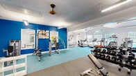 Located at 1000 McQueen Dr in Durham, NC, this fitness facility offers a comprehensive workout environment spread across a single, thoughtfully designed floor. The space is divided into two distinct exercise rooms, each serving a different training purpose while sharing a cohesive, clean aesthetic defined by white walls, large-format tile flooring, and teal-accented rubber fitness mats.

The main exercise room is the largest of the two, featuring soaring vaulted ceilings with ceiling fans and pendant lighting that enhance airflow and visibility. This area is equipped with a wide array of cardio machines — including treadmills, ellipticals, and stationary bikes — alongside selectorized strength machines and a dumbbell rack. A wall-mounted flat-screen TV and motivational artwork add to the energizing atmosphere. Large windows and a glass door allow natural light to pour in, offering views of an outdoor pool area.

The secondary exercise room provides a more intimate setting ideal for functional and flexibility training. It features full-length wall mirrors, a rack of medicine balls, resistance training equipment, and open floor space for stretching or mat work. A ceiling fan keeps the room comfortable during use.

Throughout the facility, bright fluorescent lighting ensures excellent visibility, while the crisp white interiors and accent walls in bold blue contribute to a modern, motivating environment well-suited for a variety of fitness routines.