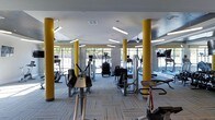 Located on 9th Street in Durham, NC, this single-floor fitness center offers a spacious, well-organized workout environment designed for functionality and comfort. The space is defined by striking yellow structural columns that add a bold visual element while reinforcing the energetic atmosphere of the room. Overhead, a combination of linear fluorescent lighting and pendant fixtures ensures the entire floor is evenly and brightly lit throughout the day.

A row of large floor-to-ceiling windows along one wall floods the interior with natural light and provides open views to the outdoors, creating an airy, inviting ambiance that enhances the workout experience. The patterned low-pile carpet tiles covering the floor offer both comfort and a clean, modern aesthetic.

The facility is outfitted with a comprehensive selection of cardio and strength training machines, including treadmills, ellipticals, stationary bikes, and various resistance equipment, all arranged to allow ample space for movement between stations. Mirrors line portions of the walls, adding depth to the room and supporting functional training. The overall design balances practicality with a polished, contemporary feel.