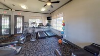 This fitness center offers a comprehensive workout environment designed to accommodate a wide range of exercise needs. The space is thoughtfully laid out across a single floor, with patterned carpet flooring throughout that defines the workout zones while adding a polished aesthetic.

Large windows line multiple walls, flooding the interior with abundant natural light and offering views of the surrounding outdoor greenery. Ceiling fans help maintain airflow throughout the room, contributing to a comfortable exercise environment. A wall of floor-to-ceiling mirrors on one side visually expands the space and supports form-checking during workouts.

The cardio section is well-stocked with multiple treadmills and elliptical machines, each positioned to face the windows for an energizing view. A wall-mounted flat-screen television is conveniently positioned for entertainment during cardio sessions.

The strength training area features a multi-station cable and weight machine, an adjustable bench, and a full rack of dumbbells spanning a range of weights. A medicine ball rack with colorful balls rounds out the functional fitness offerings. A water cooler station near the entrance ensures easy hydration access.

French-style glass doors provide entry to the space, reinforcing the refined architectural detailing seen in the crown molding and neutral wall tones. Overall, the fitness center presents a clean, functional, and inviting atmosphere suited for a variety of fitness routines.