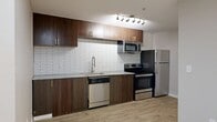 This studio is a thoughtfully designed single-floor unit that makes efficient use of its compact footprint. Light wood-style plank flooring runs continuously throughout the space, lending a warm, cohesive aesthetic to every area.

The kitchen is a standout feature, outfitted with rich dark wood cabinetry accented by gold-toned hardware, granite countertops, and a classic white subway tile backsplash. A full suite of stainless steel appliances — including a refrigerator, electric range, built-in microwave, and dishwasher — provides modern functionality. Track lighting above the kitchen adds focused illumination to the work area.

The open living area is bright and clean, with neutral gray walls, recessed lighting, and a window that allows natural light to filter in. The space offers flexibility for a variety of furniture arrangements. An electrical panel is discretely positioned along one wall.

The bathroom features a vessel-style sink set on a granite vanity top, a framed mirror, and matching wood-look flooring that ties into the rest of the unit. The angled ceiling adds an architectural detail to the space.

Overall, this studio presents a modern, well-finished interior with consistent design elements throughout, offering a functional and stylish living environment.