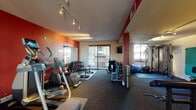 Located at 2383 N Main St in Walnut Creek, California, this fitness center is a well-equipped, single-floor facility designed to support a comprehensive workout experience. The space features bold red and orange accent walls offset by neutral tones, creating an energizing atmosphere throughout. Track lighting runs along the ceiling, ensuring the entire floor area is well-illuminated.

The main workout area is generously sized and houses a wide variety of equipment, including treadmills, elliptical trainers, a stationary bike, and a rowing machine for cardiovascular training. A dedicated free weights section offers a full dumbbell rack with a weight bench and a large wall mirror, allowing users to monitor their form. Kettlebells of varying weights are neatly arranged on a rack near a sliding glass door, which opens to an outdoor balcony area, bringing in natural light and views of the surrounding property.

A separate adjoining room provides additional space for strength training, featuring a cable resistance machine and a wall-mounted exercise mat, along with access to restroom facilities. Stability balls and open floor space throughout accommodate stretching and functional fitness routines. The durable carpet flooring and structural columns with red accents contribute to the gym's distinctive, motivating aesthetic, making this fitness center a practical and visually dynamic amenity.