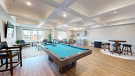 Located at 60 Beard Sawmill Road in Shelton, Connecticut, this thoughtfully designed single-floor community lounge offers a refined yet inviting atmosphere ideal for socializing and recreation. The space is immediately distinguished by its striking coffered ceiling adorned with detailed crown molding, complemented by recessed lighting that creates a bright, welcoming ambiance throughout.

Light hardwood flooring runs seamlessly across the entire space, tying together several distinct functional zones. At the center, a substantial billiards table with a vivid blue felt surface serves as the room's focal point, accompanied by a wall-mounted cue rack and a flat-screen television for entertainment. Adjacent to the billiards area, a comfortable lounge zone features quilted leather seating arranged around a coffee table, perfect for casual conversation.

A dedicated dining area provides a glass-top round table with seating for four, positioned near large windows that allow generous natural light to pour in. A wet bar with grey cabinetry, a tile backsplash, and counter seating adds a functional and stylish amenity for entertaining. A high-top pub table with bar stools occupies a cozy corner near French doors that open to an outdoor area, further extending the social space. Multiple windows throughout ensure the room remains airy and connected to the outdoors.