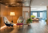 This thoughtfully designed single-floor space seamlessly integrates a variety of functional zones through a cohesive design language rooted in warm wood tones, natural materials, and curated artwork. The open-plan layout connects a spacious corridor, kitchen, lounge, and dining areas, unified by wide-plank hardwood flooring and large-format tile that transitions fluidly between zones.

Vertical wood slat partitions serve as elegant dividers throughout, offering visual separation without sacrificing openness. The kitchen features a generous waterfall-edge island with bar seating, warm wood cabinetry, stainless steel appliances, and a striking skylight overhead that floods the space with natural light.

A bold teal accent wall defines the living area, where a plush seating arrangement centers around a large-screen television mounted on a textured wall panel. The most dramatic feature is a vivid illustrated ceiling mural depicting botanicals and figurative art, framed by a coffered tray ceiling with ambient lighting—adding artful character to both the lounge and dining room.

The dining area accommodates a full-sized conference-style table surrounded by contemporary chairs, with large windows drawing in city views. Large-scale photographic artwork and sculptural accents throughout reinforce a gallery-like atmosphere, making this a refined and visually dynamic space.